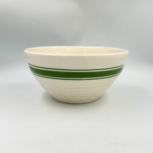 Puede incluir: Un cuenco de cerámica de color crema con una franja horizontal verde ancha en el centro, acentuada por dos líneas verdes más finas arriba y abajo, con sutiles estrías horizontales a lo largo de la superficie exterior inferior.