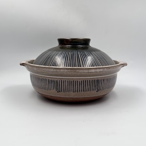 May include: A ceramic cooking pot with a lid. The pot is beige with vertical black stripes around the body and lid. The lid has a dark brown knob. The pot has two small handles on either side.