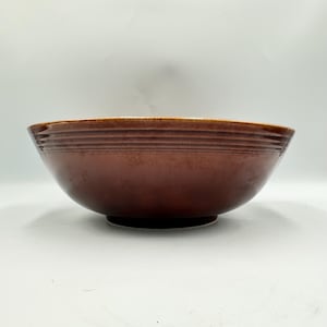 May include: A brown ceramic bowl with a wide, shallow design. The bowl has a dark brown exterior with horizontal ridges near the rim. The interior is a lighter shade of brown. The bowl sits on a small, circular base.