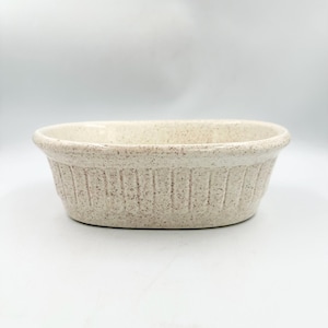 May include: An oval-shaped ceramic baking dish with a speckled cream and tan finish. The exterior features a series of vertical fluted ridges around the sides, while the interior is smooth with a wide, rounded rim along the top edge.