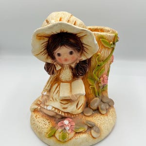 Puede incluir: Figura de cerámica de una niña con un sombrero grande, leyendo un libro. La niña está sentada junto a un jarrón con forma de tronco de árbol, adornado con flores y enredaderas. La paleta de colores incluye beige, marrón y rosa.