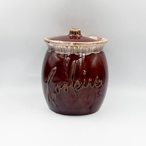 May include: A round, dark brown ceramic cookie jar with a matching lid. The jar features a glossy finish and a textured, cream-colored drip glaze around the rim. The word Cookies is embossed in a cursive script on the front of the jar body.