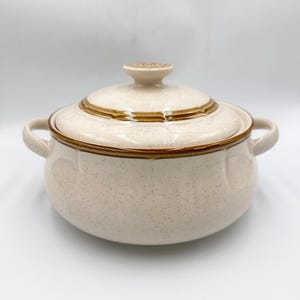 May include: A beige ceramic casserole dish with a lid. The dish has two handles and a brown trim around the rim and lid. The lid has a small knob on top. The dish has a speckled pattern.