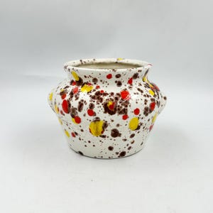 May include: A small, wide-mouthed ceramic pot with a white base featuring a speckled pattern of dark brown, bright yellow, and vibrant red spots. The vessel has a rounded body, a slightly flared rim, and two small, integrated side handles.
