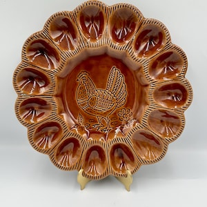 May include: A brown ceramic egg serving platter with a scalloped edge and a central rooster design. The platter has 12 egg-shaped indentations around the perimeter. The rooster is etched into the center of the platter.