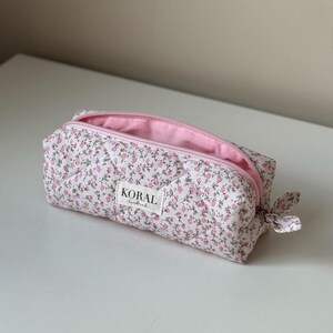Op de afbeelding: Een rechthoekige etui met bloemenpatroon. De witte stof is bedrukt met kleine roze bloemen. De binnenkant en rits zijn roze. Het label luidt "KORAL handmade". Een klein strikje is bevestigd.