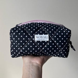 Puede incluir: Un estuche acolchado negro con un patrón de lunares blancos y una cremallera de color rosa claro. Una etiqueta de tela rectangular blanca está centrada en la parte frontal con el texto negro KORAL handmade impreso en una fuente sans-serif.