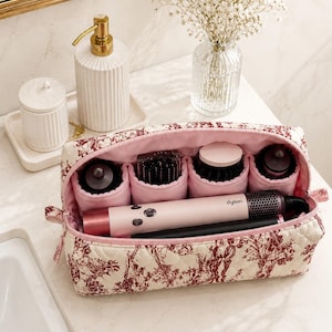 Può includere: Una borsa da toilette rettangolare trapuntata con un motivo floreale rosso e bianco. L'interno è foderato in rosa chiaro e contiene uno strumento per lo styling dei capelli Dyson con diversi accessori cilindrici organizzati in scomparti.