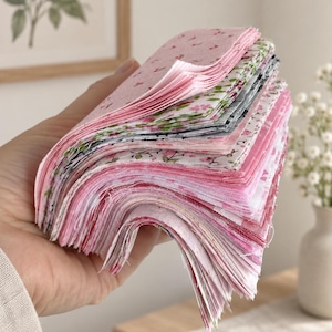 Könnte beinhalten: Ein Stapel quadratischer Stoffzuschnitte in verschiedenen Rosa-, Weiß- und Grüntönen. Die Sammlung umfasst Muster mit kleinen Blumenprints, Kirschmotiven und Vichy-Karos, die alle mit rohen, leicht ausgefransten Kanten in einheitlicher Größe geschnitten sind.