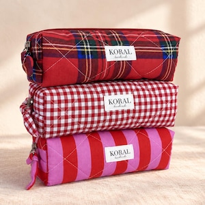 May include: A stack of three rectangular quilted pencil cases with zipper closures. The top case features a red, green, and blue plaid pattern. The middle case has a red and white gingham check. The bottom case has red and pink vertical stripes.
