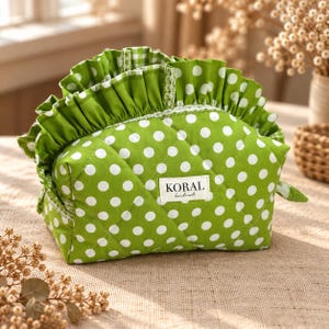 May include: A quilted green pouch with white polka dots, featuring a ruffled top edge and a white fabric label centred on the front that reads KORAL handmade. The pouch has a structured, rounded shape with visible diamond-patterned stitching.