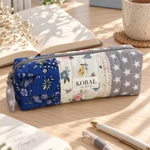 Pode incluir: Um estojo acolchoado retangular com um design de patchwork de tecido floral azul, tecido floral creme, uma tira de renda branca e tecido cinza com estrelas brancas. Uma etiqueta diz KORAL handmade. Possui um zíper cinza com um puxador de estrela.
