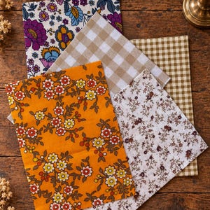 Op de afbeelding: Een verzameling van vier rechthoekige stoffen met verschillende patronen: één met een levendige oranje achtergrond en veelkleurige bloemenprint, één met een witte achtergrond en kleine donkere bloemtakjes, één met een beige en witte gingham ruit, en één met een witte achtergrond en een opvallend paars en blauw bloemmotief.