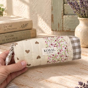 Pode incluir: Um estojo retangular acolchoado com um design de patchwork de tecido creme com corações castanhos, uma estampa floral, acabamento em renda branca e uma extremidade em xadrez cinza e branco. Uma etiqueta branca diz KORAL handmade.