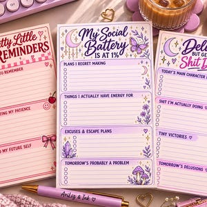 May include: Three rectangular paper notepads with printed headers and checklists. The left pad is pink with cherry and bow graphics titled Petty Little Reminders. The center pad is purple with moon and mushroom art titled My Social Battery Is At 1%. The right pad is purple with butterfly art titled Delulu But Getting Shit Done.