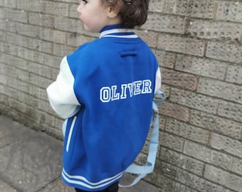 Op de afbeelding: Een blauw en wit baseballjack met de naam "OLIVER" geborduurd op de rug. Het jack heeft witte mouwen en een blauw lichaam met witte strepen rond de kraag en manchetten.