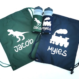 Op de afbeelding: Twee trekkoordtassen, een groene en een blauwe, met de gepersonaliseerde namen "Jacob" en "Myles". De groene tas heeft een wit dinosaurus silhouet, terwijl de blauwe tas een wit treinsilhouet heeft. Twee waterflessen met bijpassende dinosaurus- en treindesigns zijn ook inbegrepen.