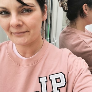 Könnte beinhalten: Ein rosa Sweatshirt mit einem weißen Filz-Patch, auf dem "JP" steht.