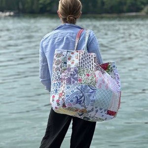 May include: A large patchwork tote bag with a variety of floral and patterned fabric squares. The bag has a white background with red, blue, and purple accents. The tote bag is carried over the shoulder.