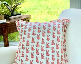 May include: A white pillow with a pink bunny rabbit print. The pillow has a ruffled edge and is sitting on a white couch.