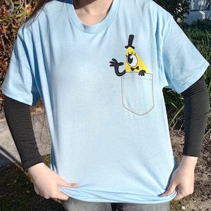 May include: Light blue t-shirt featuring a cartoon character design in the pocket. The character is a yellow triangle with a black top hat, bow tie, and one eye. The shirt has short sleeves and is worn over a black long-sleeved shirt.