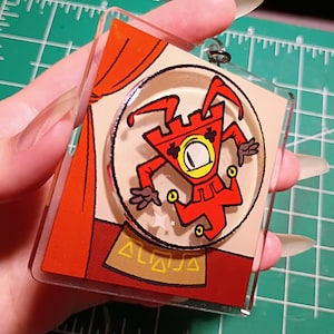 May include: A square acrylic keyring featuring a cartoon jester character in a red suit with a single large eye, positioned inside a circular crystal ball graphic. The background shows orange curtains and a gold base with geometric triangle patterns.