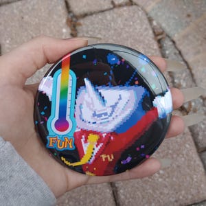 May include: A round, glossy button with a pixelated design. The button features a rainbow thermometer and the word "FUN" in yellow. The background is black with colorful confetti and a pixelated character.
