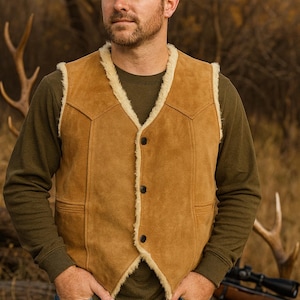 May include: A tan suede vest with cream-colored shearling trim and black buttons. The vest is worn over a green long-sleeved shirt and paired with blue jeans. A brown baseball cap completes the look.