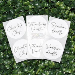 May include: White, round labels with black script text. The labels read "Chocolate Chip", "Strawberry Vanilla", and "Snicker-doodle". The labels are on a green, leafy background. The text "Vanna Customs" is also visible.