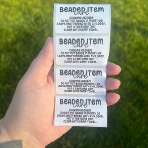 May include: A stack of white rectangular labels with black text. The labels read "BEADED ITEM care" and include a choking hazard warning, instructions for use, and cleaning instructions. The labels are held against a green background.