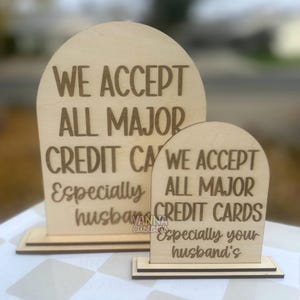 May include: Two wooden signs with arched tops and rectangular bases. The larger sign reads "WE ACCEPT ALL MAJOR CREDIT CARDS Especially husband's". The smaller sign has the same text. The signs are light brown.