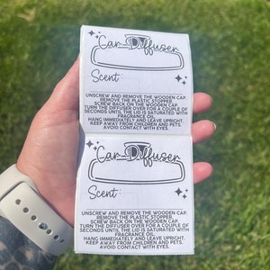 May include: Two white labels with black text and a black and white illustration of a car air freshener. The text on the label reads "Car Diffuser" and "Scent: Unscrew and remove the wooden cap. Remove the plastic stopper. Screw back on the wooden cap. Turn the diffuser over for a couple of seconds until the lid is saturated with fragrance oil. Hang immediately and leave upright. Keep away from children and pets. Avoid contact with eyes."