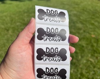 May include: A vertical strip of four rectangular adhesive labels, each featuring a black bone-shaped graphic with the white text DOG treats printed in a combination of bold block lettering and a cursive script font style.
