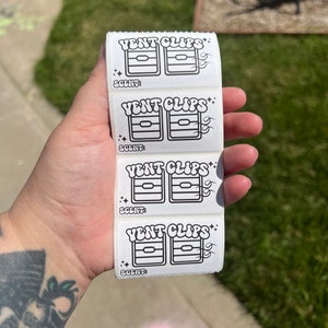 May include: A stack of white rectangular stickers with the words "VENT CLIPS" in a retro font. Each sticker features a black and white graphic of a car vent clip and the word "SCENT:" below. The stickers are held in a hand.