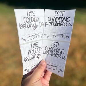 May include: White stickers with the phrases "THIS FOLDER belongs to" and "ESTE CUADERNO pertenece a" in black script. Each sticker has a ruler graphic and star accents. The stickers are held in a hand against a blurred background.
