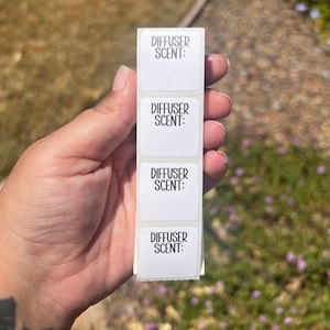 May include: A strip of white labels with the text "DIFFUSER SCENT:" printed in black. The labels are rectangular and are held against a blurred green and brown background.
