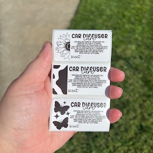 May include: Three rectangular white stickers with black text and graphics. Each label is titled Car Diffuser Care and includes instructions for use. The designs feature a sunflower, a cow print pattern, and two butterflies with stars.
