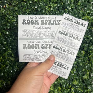 May include: A hand holds several white rectangular labels for room spray products. Each label features the text "Your Business Name," "ROOM SPRAY," and "Scent Name" in a stylized font. The labels are against a backdrop of green foliage.