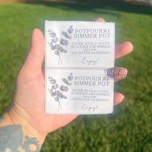 May include: Two white rectangular labels with text for a potpourri simmer pot. The labels have a floral design and the text "POTPOURRI SIMMER POT", instructions, and the word "Enjoy!". The labels are held in a hand.