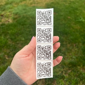 May include: Four white square stickers with black QR codes printed on them. The stickers are arranged vertically and are held in a person's hand.
