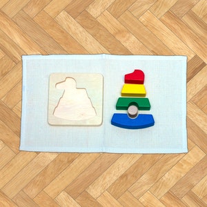 May include: A wooden stacking toy with five colorful rings in red, yellow, green, blue, and brown. The rings are stacked on a wooden base. The toy is on a white mat.
