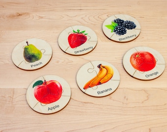 May include: Six wooden puzzle pieces with images of different fruits and a blackberry. Each piece has a different fruit image: a peach, a strawberry, a banana, an apple, an orange, and a blackberry. The pieces are labeled with the name of the fruit.
