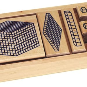 May include: A wooden Montessori math set with various blocks and shapes. The set includes a large cube, a flat square, a long rectangular block, and several smaller cubes, all with dark blue grid patterns. The blocks are contained within a light-colored wooden tray.