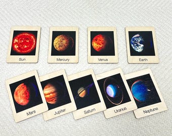 Pode incluir: Um conjunto de nove cartas de madeira com imagens dos planetas do nosso sistema solar, incluindo o Sol. Cada carta tem um fundo preto e um rótulo branco com o nome do planeta impresso nela.
