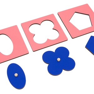 May include: A set of pink and blue wooden shape puzzle pieces. The pink pieces have cutouts of an oval, a four-leaf clover, and a pentagon. The blue pieces are solid shapes that fit into the cutouts.