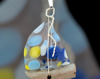 May include: A handcrafted sailboat pendant with a clear sail adorned with blue and yellow glass accents. The sail is attached to a driftwood base and suspended by a silver-toned metal wire. The pendant is designed to be worn as a necklace.