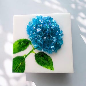 May include: Square white canvas art featuring a blue hydrangea flower made of sparkling, crushed glass. The flower has a detailed green stem and leaves. The artwork is a unique, handcrafted piece.