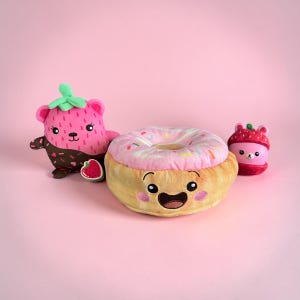 May include: A plush donut with a pink frosted top and colorful sprinkles, featuring an embroidered face with a wide smile. Beside it sits a pink strawberry-themed plush bear with chocolate-dipped paws, a small strawberry charm, and a small strawberry bear toy.