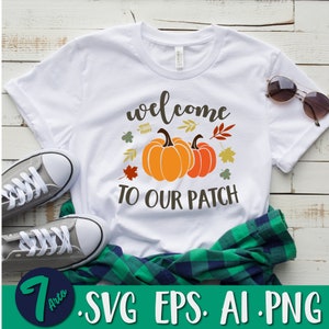 Op de afbeelding: Wit T-shirt met de tekst "Welkom op onze patch" en twee oranje pompoenen met groene stelen en bladeren. Er zijn ook herfstbladeren in verschillende kleuren rond de pompoenen.