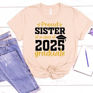 May include: A beige t-shirt with the text "Proud Sister of a class of 2025 graduate" in gold and black lettering. The shirt is laid out on a wooden surface with a pair of jeans and a notebook.